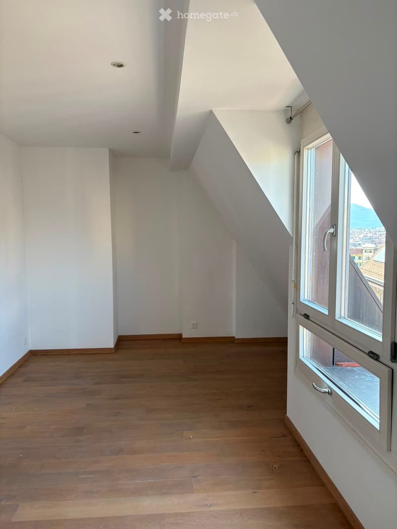 Empty attic room with wooden flooring, white walls, and a large window offering a view of the city.