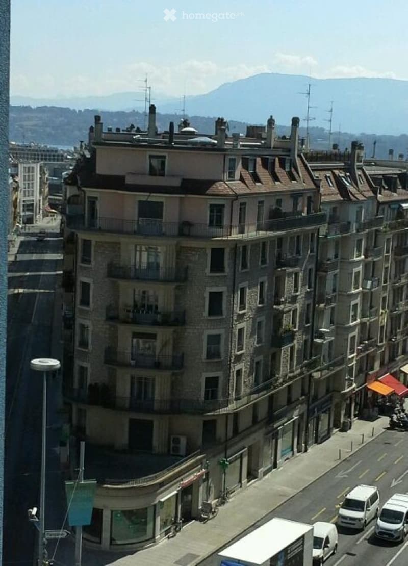Multi-story urban building with balconies and chimneys, overlooking a city street and distant mountains.