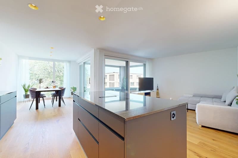 Bright, open-plan living and dining area with wooden flooring, large windows, a modern kitchen island, a sofa, a dining table with chairs, and a television.