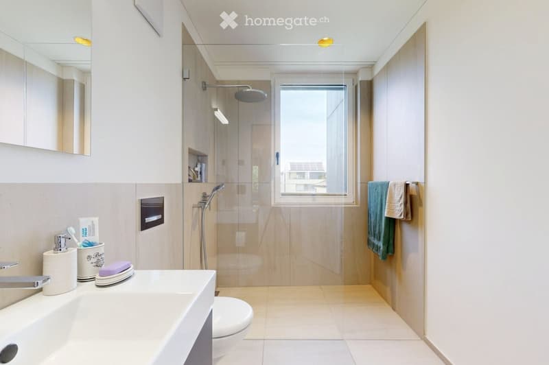 Modern bathroom with a large window, a walk-in shower, a sink with a modern faucet, a toilet, and a mirror. The space features light-colored tiles and a sleek design.