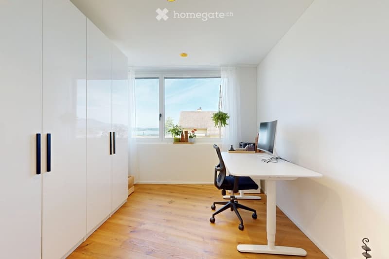 Bright, modern room with wooden flooring, large window with view, white desk, ergonomic chair, computer monitor, and wardrobe.