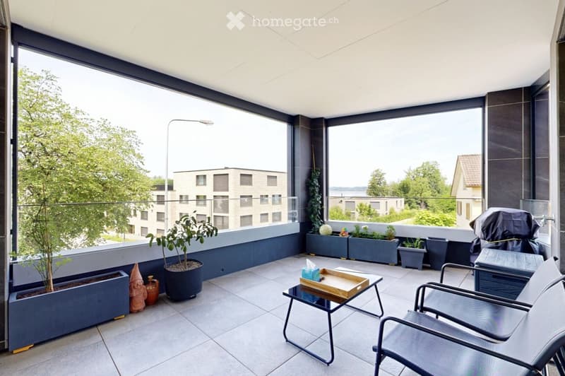 Modern balcony with large windows, tiled floor, potted plants, and outdoor furniture including chairs and a small table.