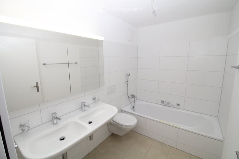 Modern bathroom with white tiles, a double sink, a toilet, a bathtub, and a large mirror above the sink.