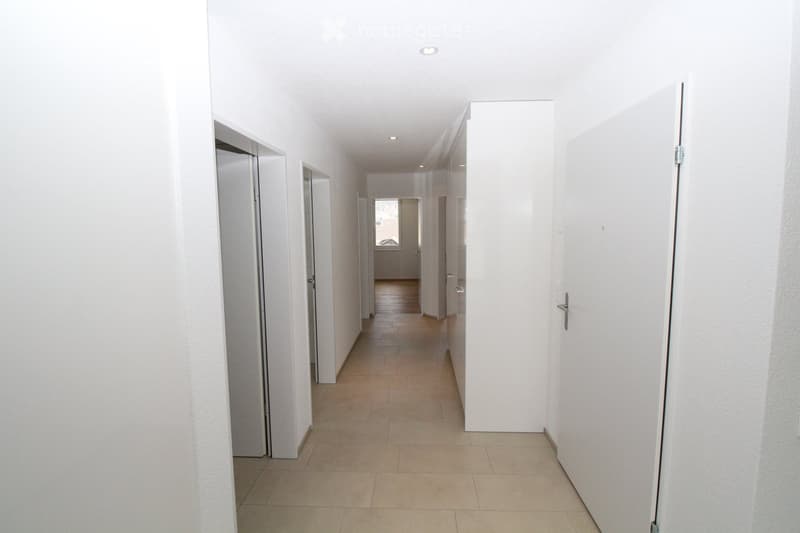 Empty hallway with white walls, tiled floor, and several closed doors on both sides.