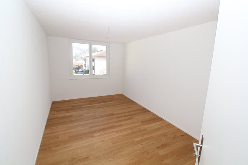 Empty room with light wooden flooring and white walls, featuring a large window with a view of the outside.