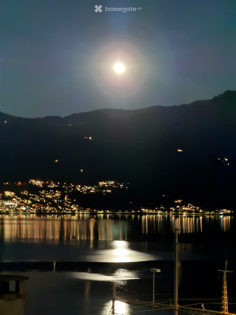 Nighttime view of a calm lake reflecting the moon and city lights from a distant town on the shore.