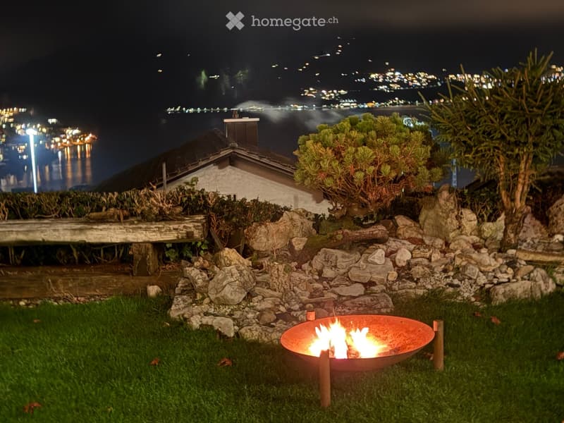 Nighttime garden with a fire pit, stone rocks, greenery, and a cityscape view in the background.