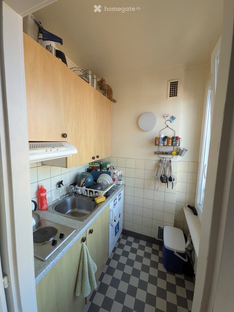 Compact kitchen with light wooden cabinets, a sink, stove, and various kitchen items on the counter and shelves.