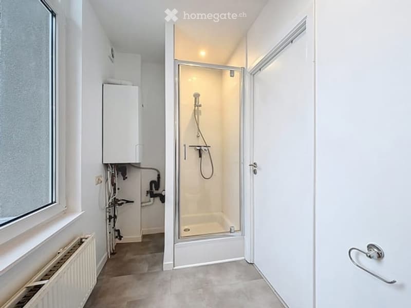 Modern bathroom with a glass-enclosed shower stall, radiator, and water heater unit visible. The space features white tiled walls and a grey tiled floor.
