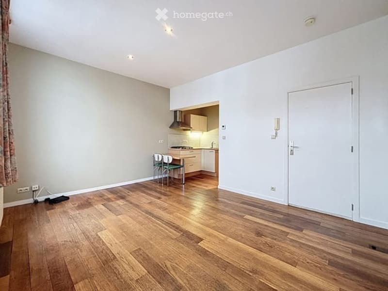 Bright, open-plan living and dining area with wooden flooring, white walls, and a small kitchen area featuring a stove, range hood, and cabinets. A dining table with chairs is positioned near the kitchen.