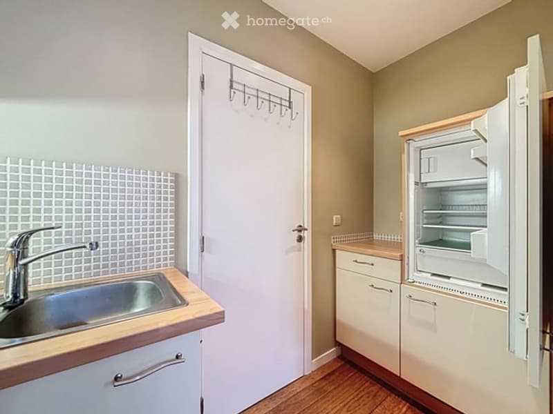 Modern kitchen with light-colored cabinets, a stainless steel sink, and an open refrigerator. The space features wooden flooring and a tiled backsplash.