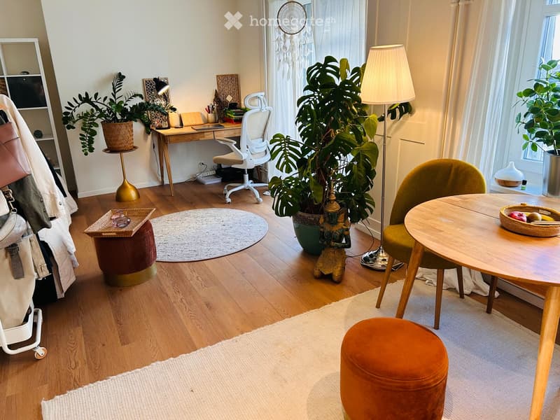 A cozy, well-lit room with wooden flooring, a round table with a green chair, a small desk with a chair, a large plant, and decorative items including a dreamcatcher and a small statue.