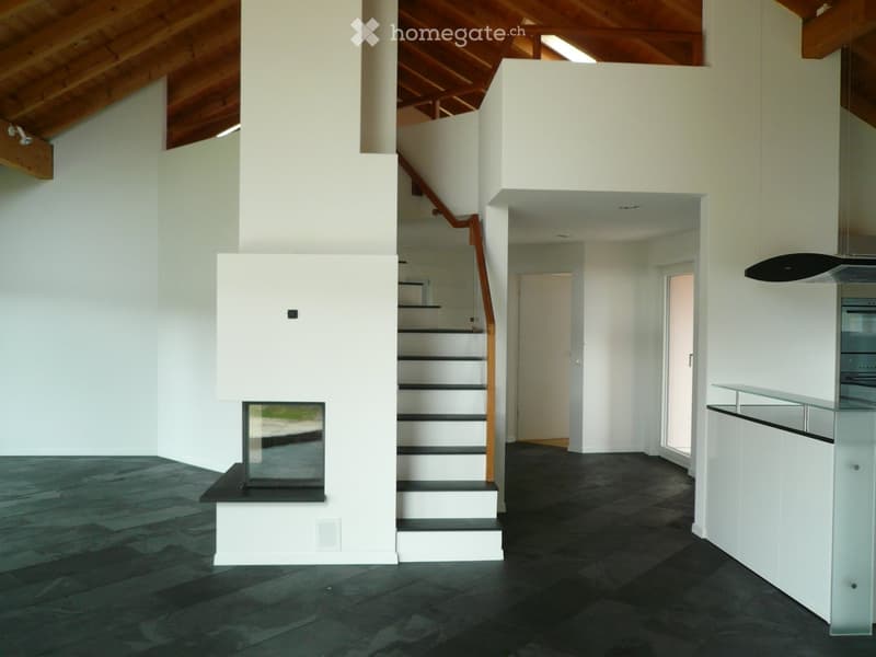 Open-plan space with a raised ground floor, wooden staircase, fireplace, and modern kitchen area. The room features dark flooring and white walls.