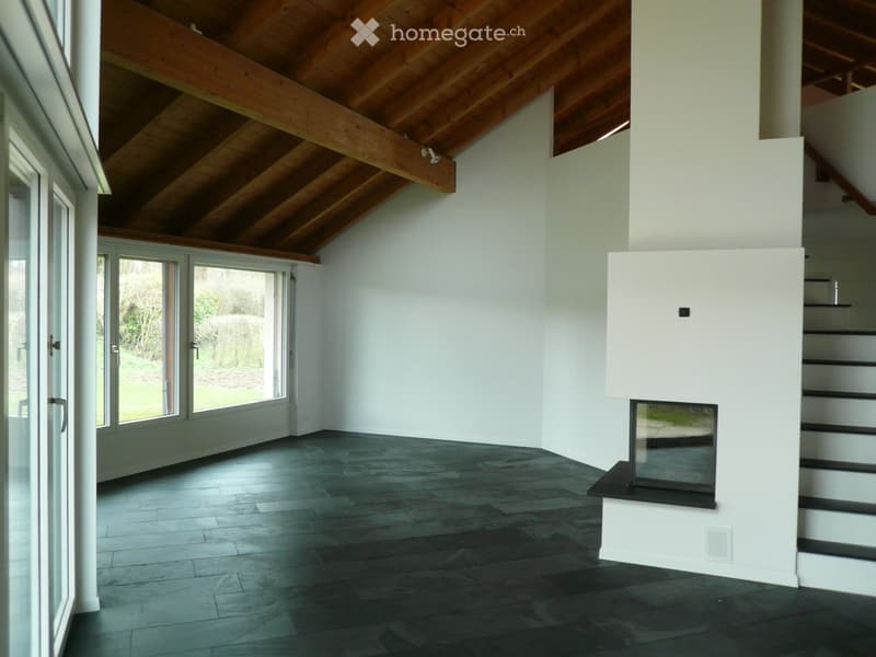 Bright, open living space with high wooden-beamed ceiling, dark hardwood floor, large glass doors leading to outdoor area, and a modern white fireplace with black surround.