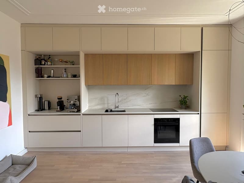 Modern kitchen with light wood cabinets, marble countertop, stainless steel appliances, and a small dining area with a chair and table.