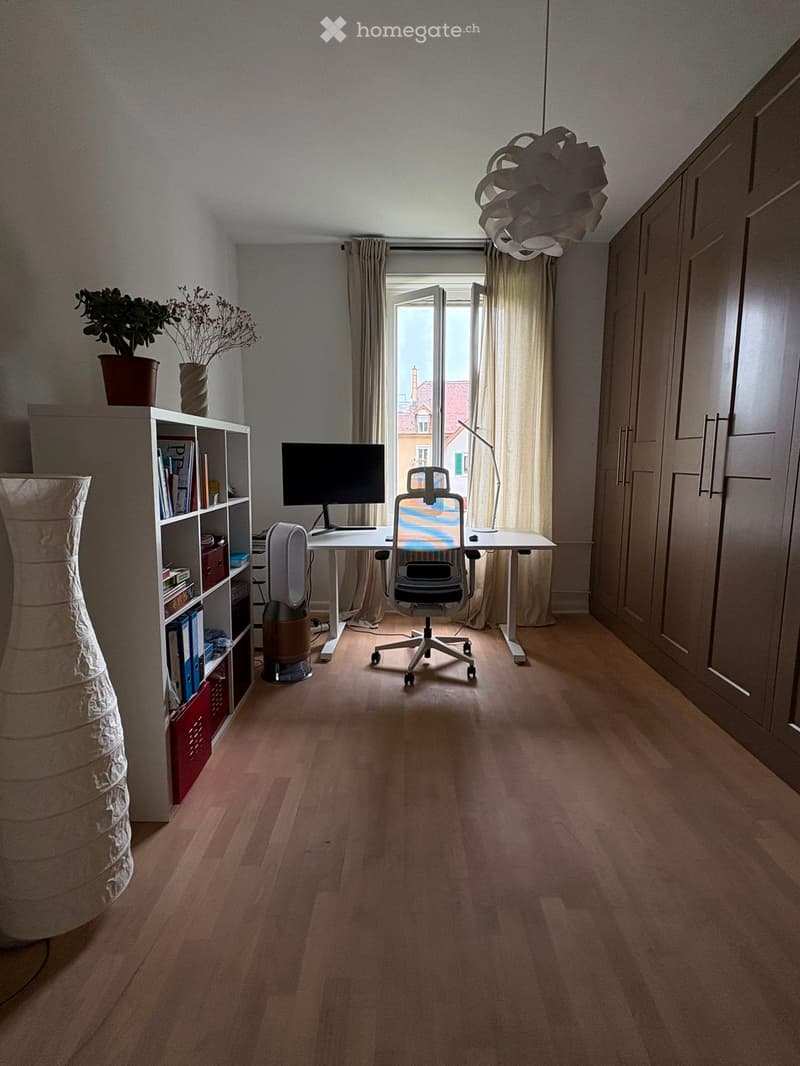 Bright, modern room with wooden flooring, large window with open curtains, desk with computer and chair, bookshelf, and decorative items.