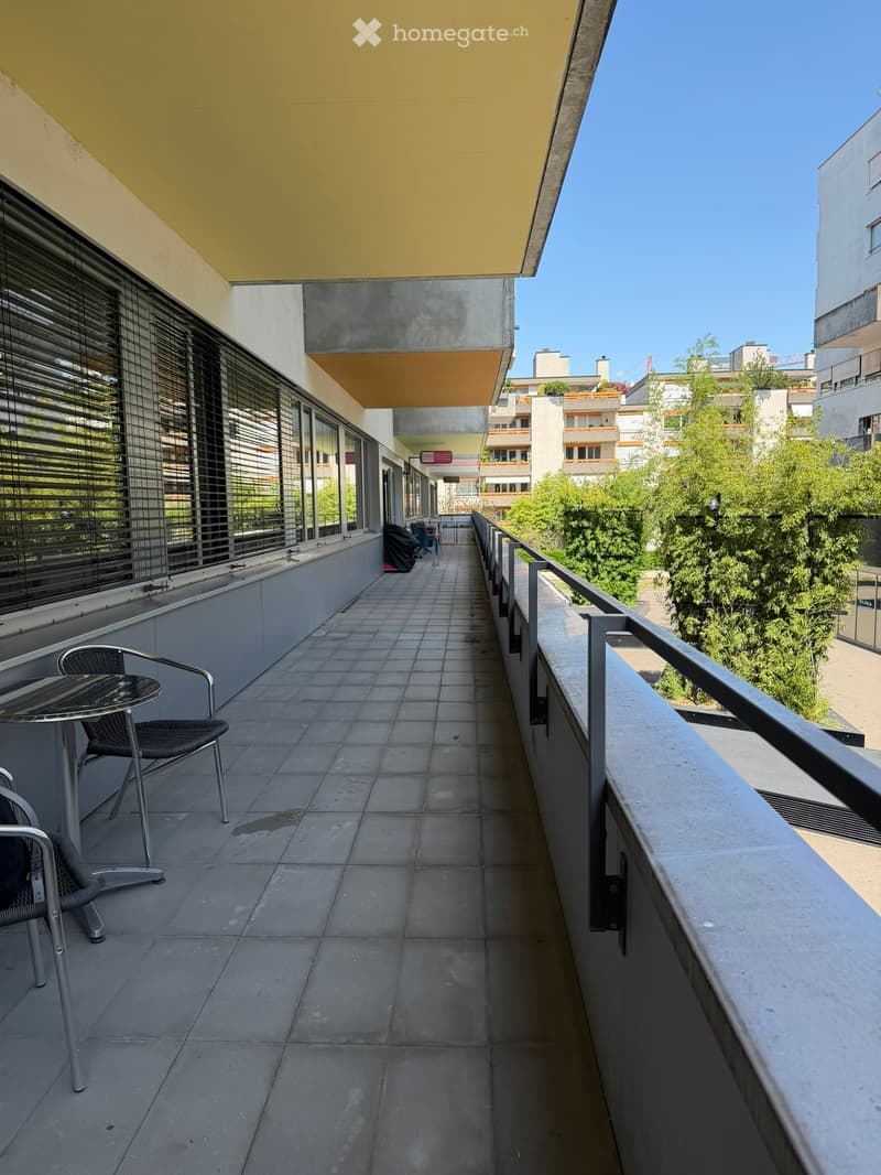 Balcony with tiled floor, metal table and chairs, metal railing, and a view of surrounding buildings and greenery.