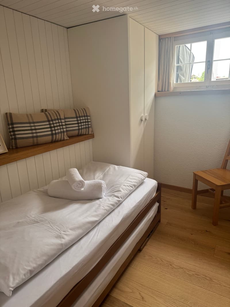 Cozy bedroom with a single bed, neatly made with white sheets and pillows, a wooden shelf with folded towels, a small wooden chair, and a window with curtains.