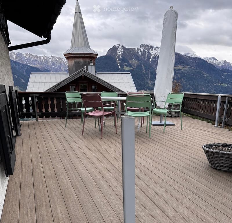 Wooden balcony with a table and chairs, overlooking a mountain landscape with a church spire visible in the distance.