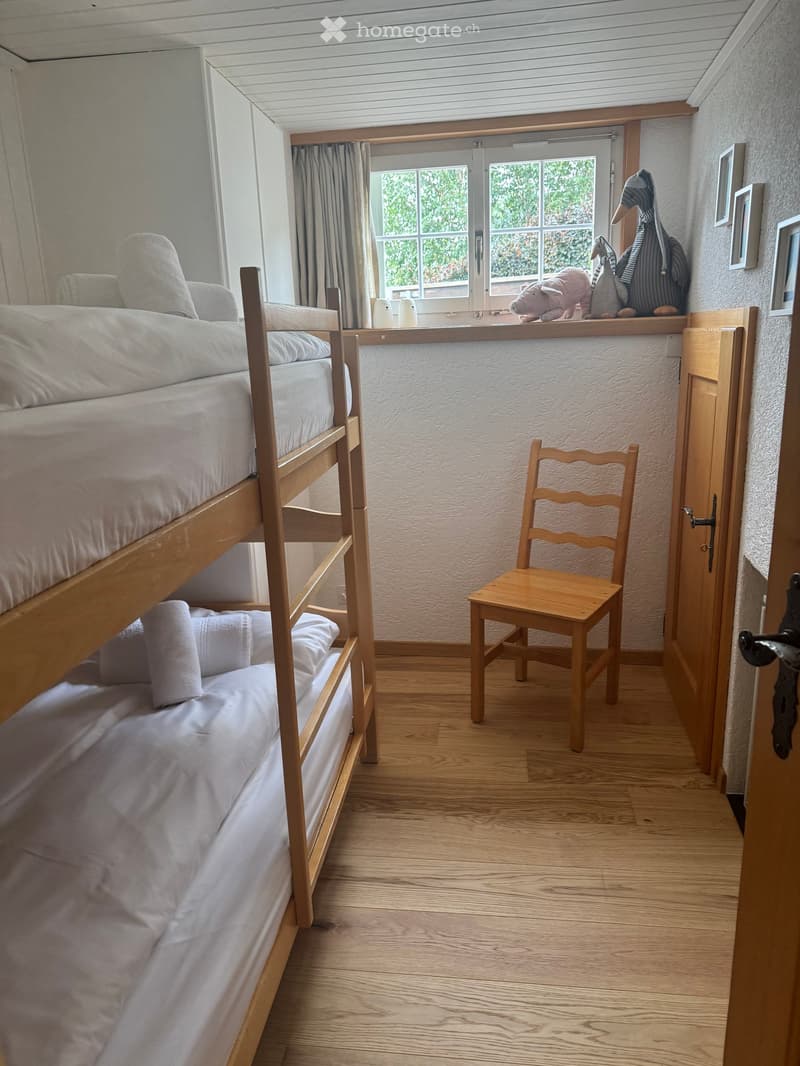 Cozy bedroom with a wooden bunk bed, a wooden chair, and decorative toys on the windowsill. Light-colored bedding and wooden flooring.
