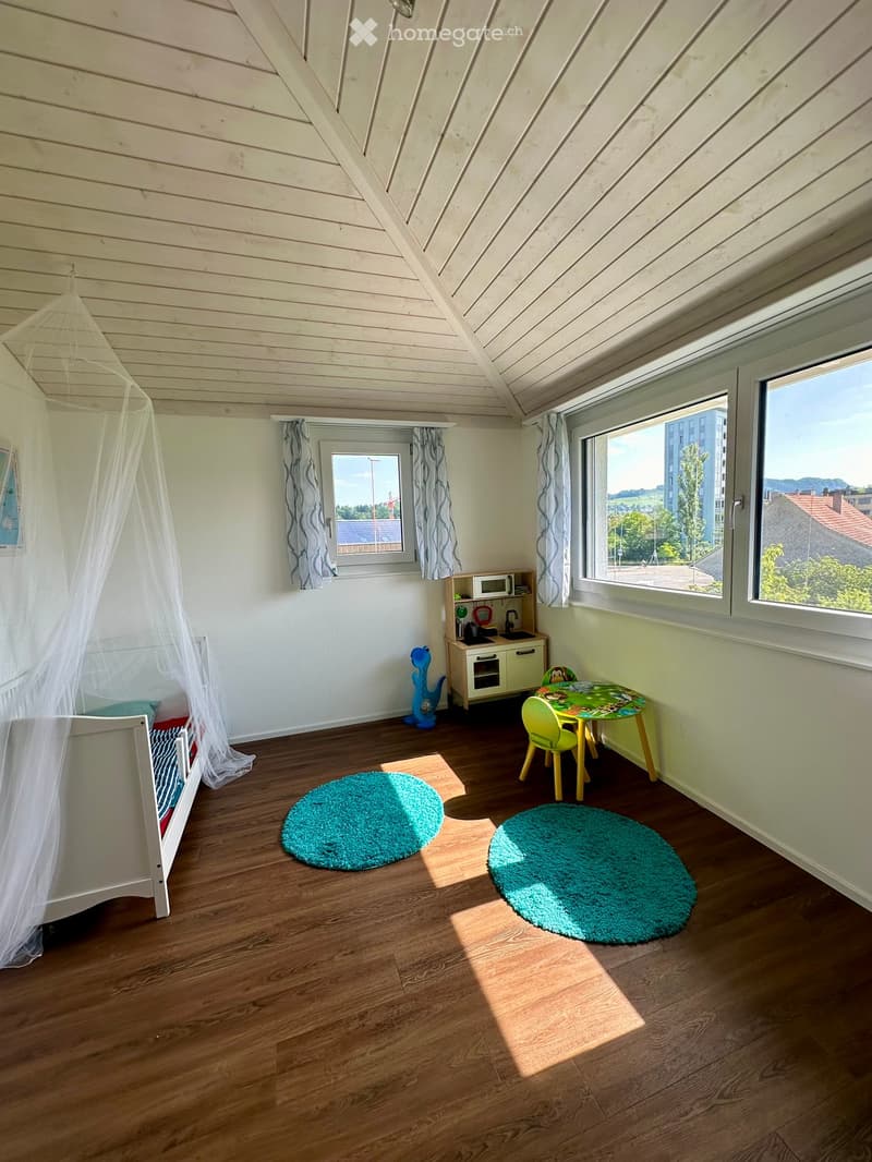 Brightly lit room with wooden floors, large windows, crib, small table and chairs, microwave, sink, and toys.