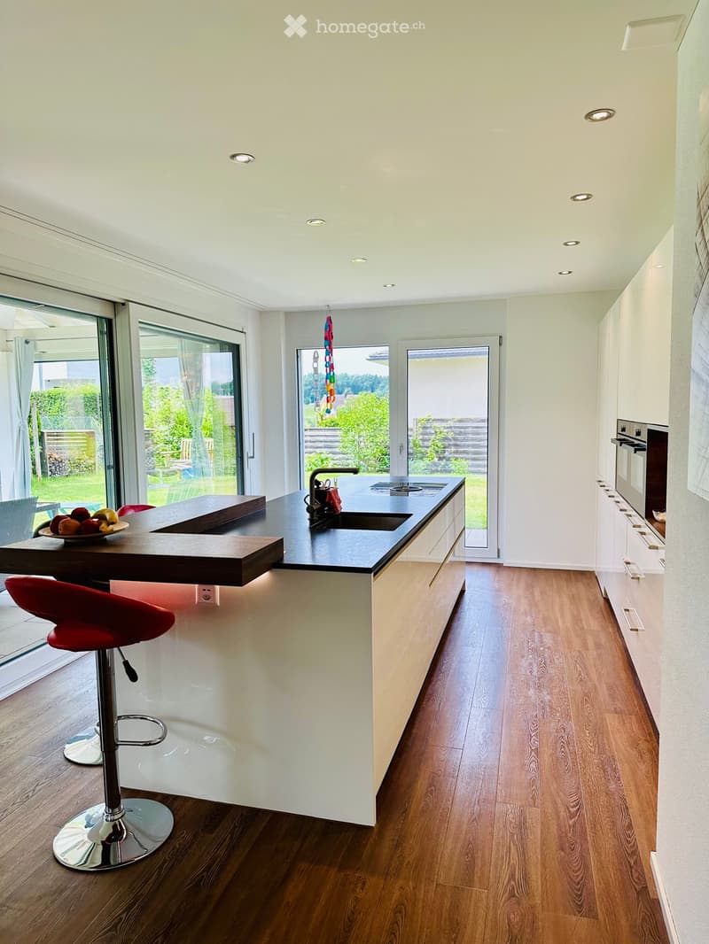 Modern kitchen with white cabinets, dark countertops, wooden flooring, and large windows overlooking a garden. Features a central island with a sink, a red stool, and a bowl of fruit on the counter.