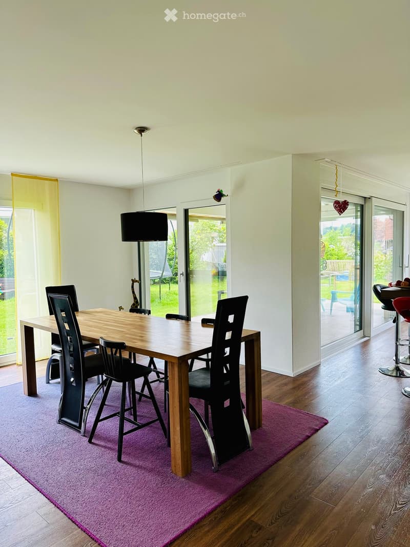Dining room with a wooden dining table, black chairs, a purple rug, and a hanging lamp. Large windows and sliding glass doors lead to a garden area.