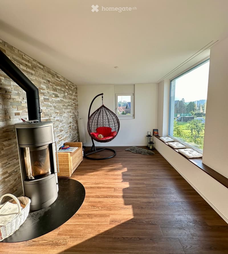 Modern living room with a stone fireplace, a hanging wicker chair, a wicker basket, and large windows offering a scenic view.