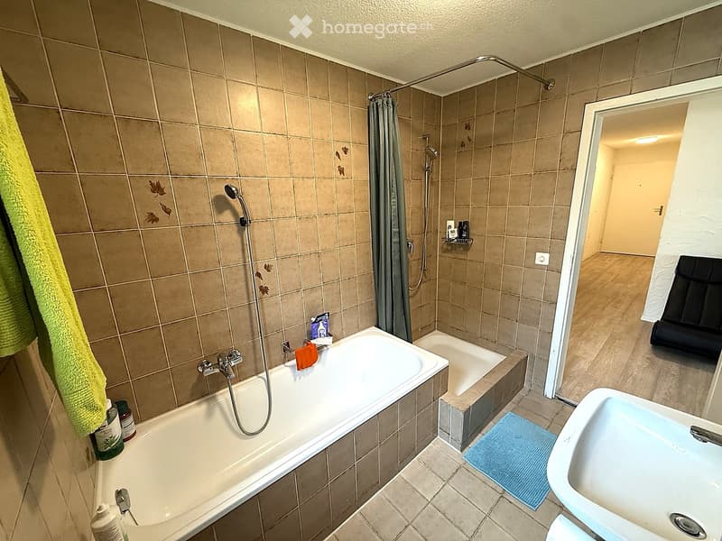 Bathroom with a bathtub, shower area, sink, and various toiletries. Beige tiled walls and floor, with a green towel hanging on the left side.