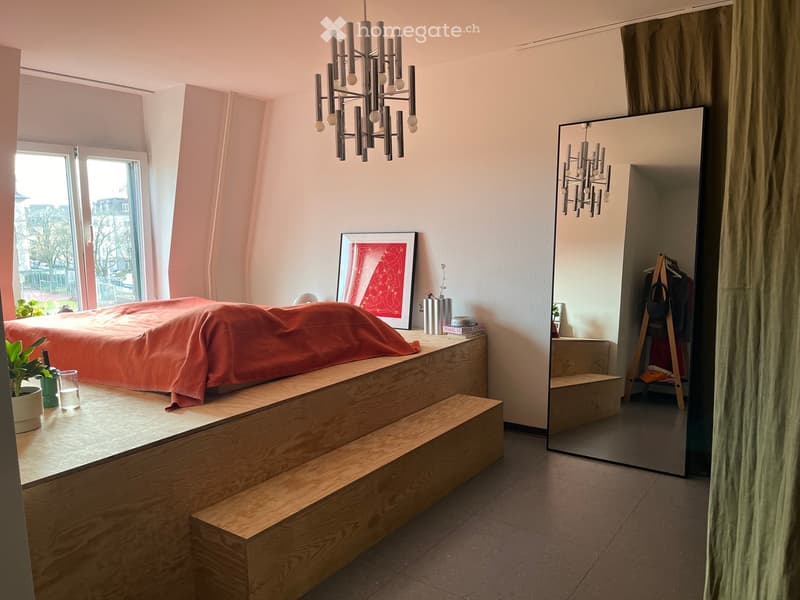 Modern bedroom with a raised wooden bed platform, large mirror, chandelier, and decorative items.