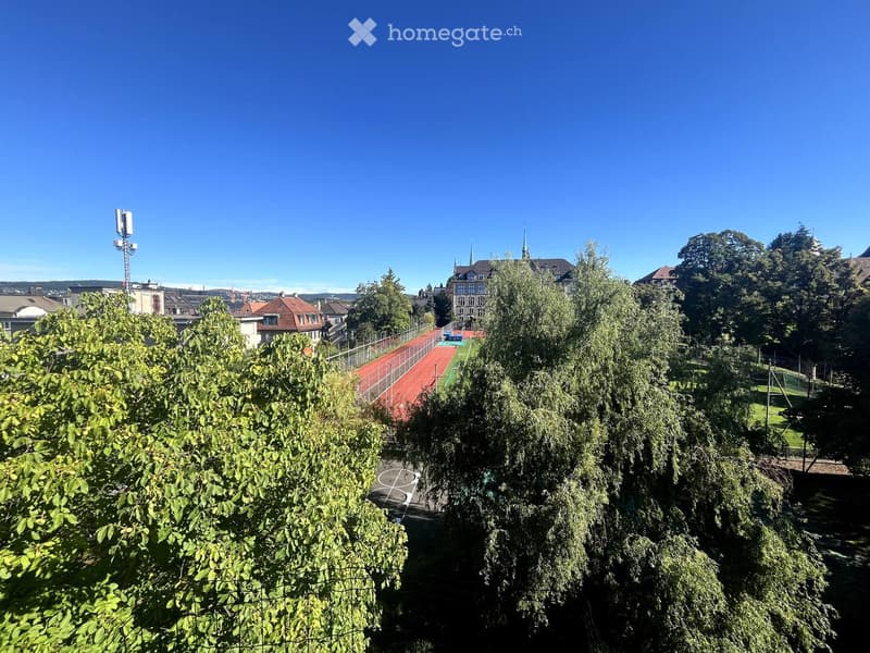 View of a park area with trees, a running track, and a sports field surrounded by residential buildings and a clear blue sky.