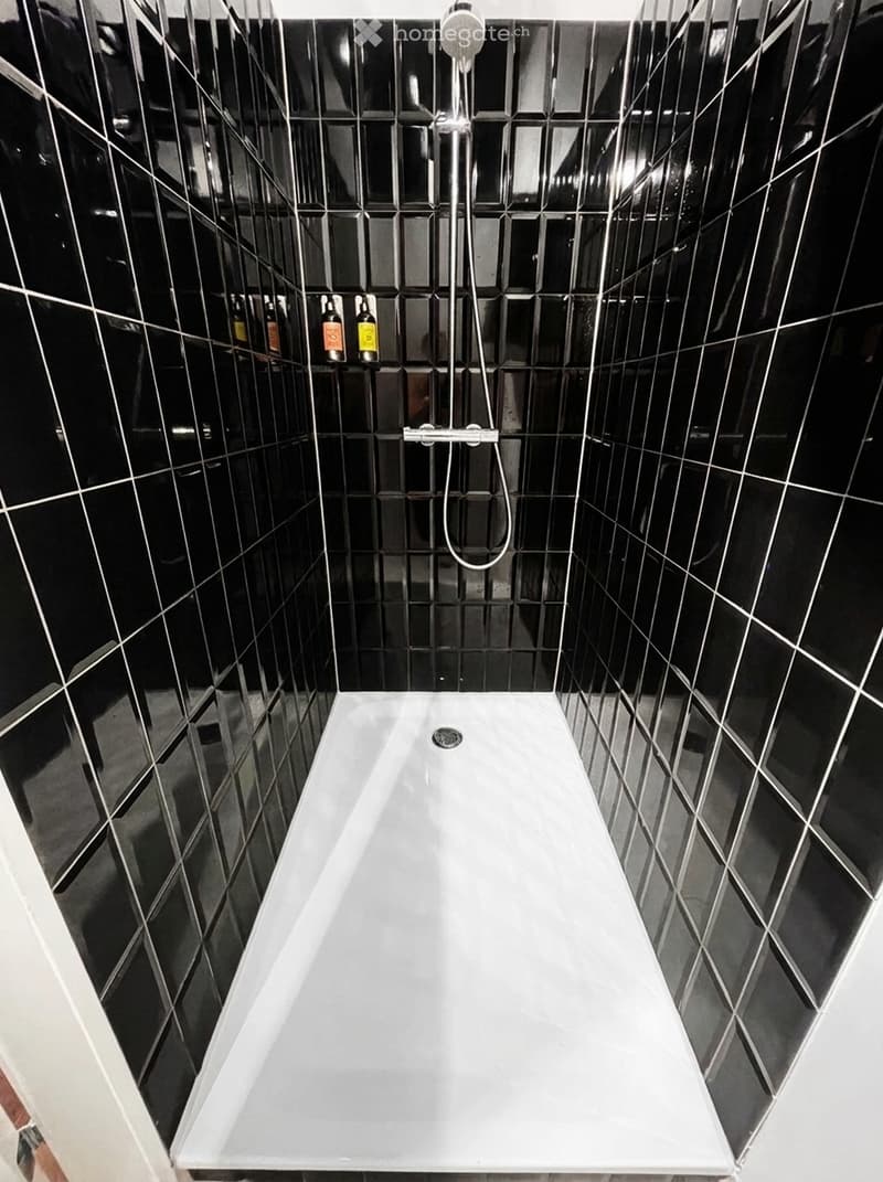 Modern bathroom with a white bathtub, black tiled walls, and a showerhead with a hose mounted on the wall. A small shower rack holds several bottles.