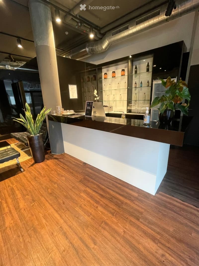 Modern reception area with a sleek black and white reception desk, laptop, two stools, potted plants, and shelves displaying various bottles.