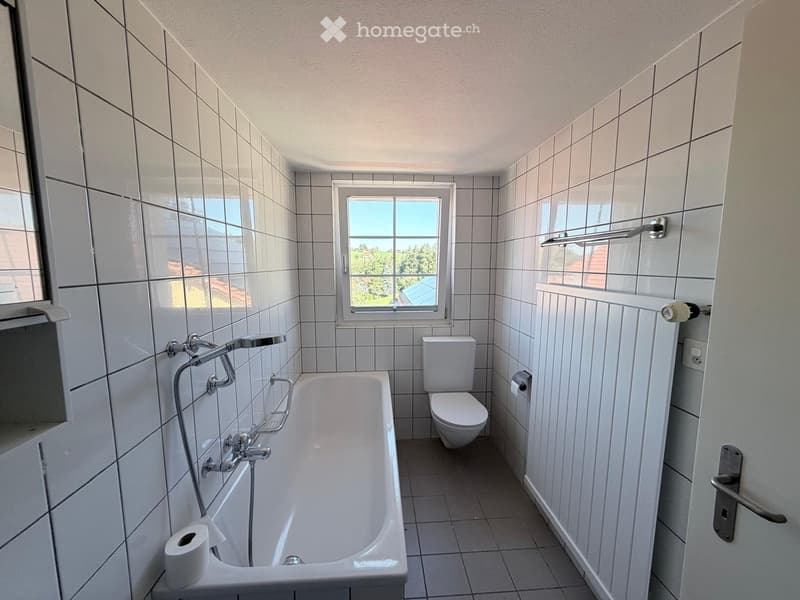Bathroom with white tiled walls and floor, bathtub, toilet, and a window offering a view of greenery outside.