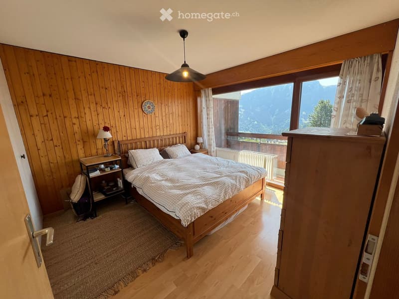 Cozy bedroom with wooden walls and floors, large window offering a scenic mountain view, a neatly made bed, nightstand, and a small table with a lamp.