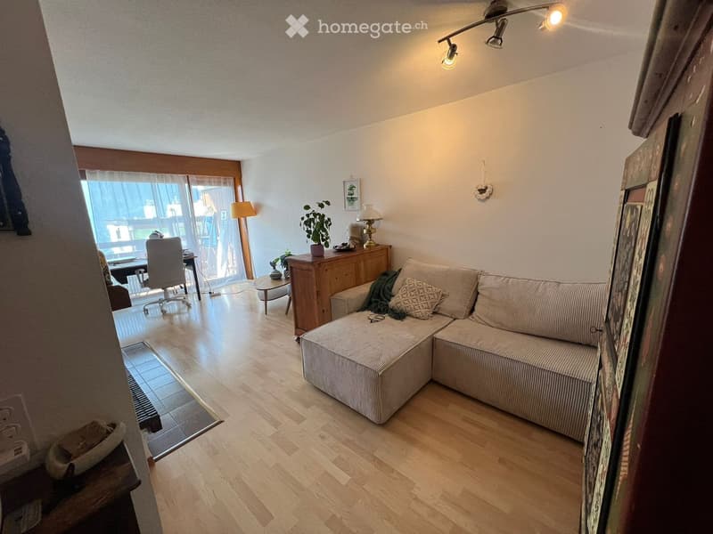 Modern living room with light wooden flooring, a sectional sofa, coffee table, side table, and a view through large glass doors.