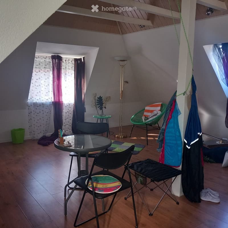 A cozy, well-lit room with wooden flooring, a round glass table, several chairs, a floor lamp, and a window with curtains.