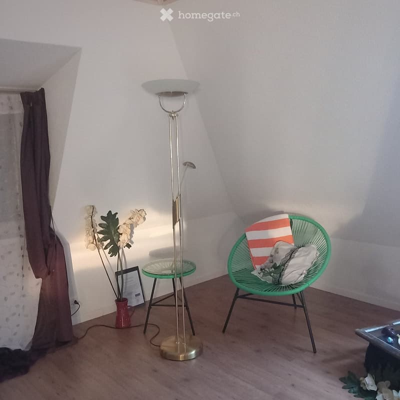 A small, cozy room with a green wicker chair, a glass side table, a floor lamp, a vase with flowers, and a picture frame on the floor.