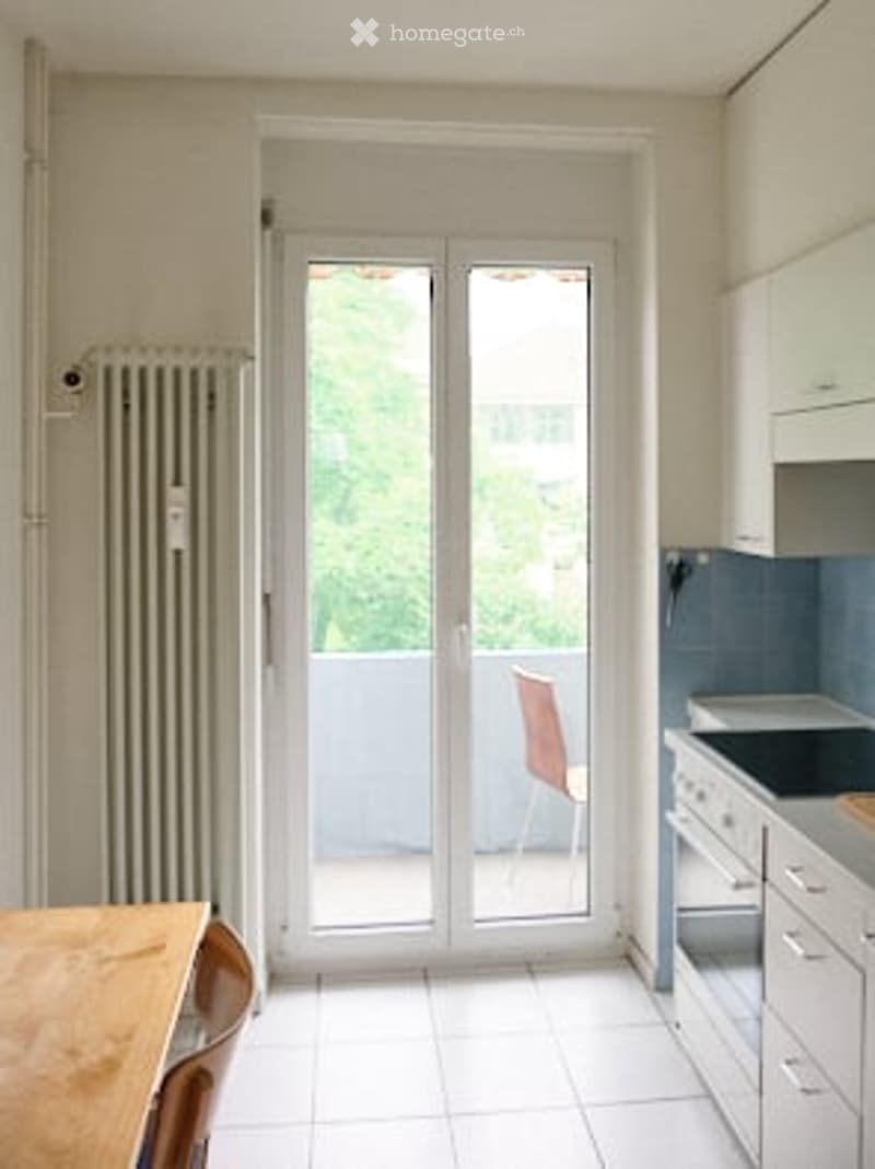 Bright, modern room with white tiled flooring, large sliding glass doors leading to a balcony, a wooden dining table with chairs, a radiator, and a kitchen area with cabinets and an oven.