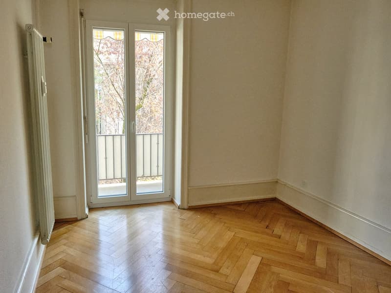 Empty room with wooden parquet flooring, light-colored walls, and a large glass door leading to a balcony with a metal railing.