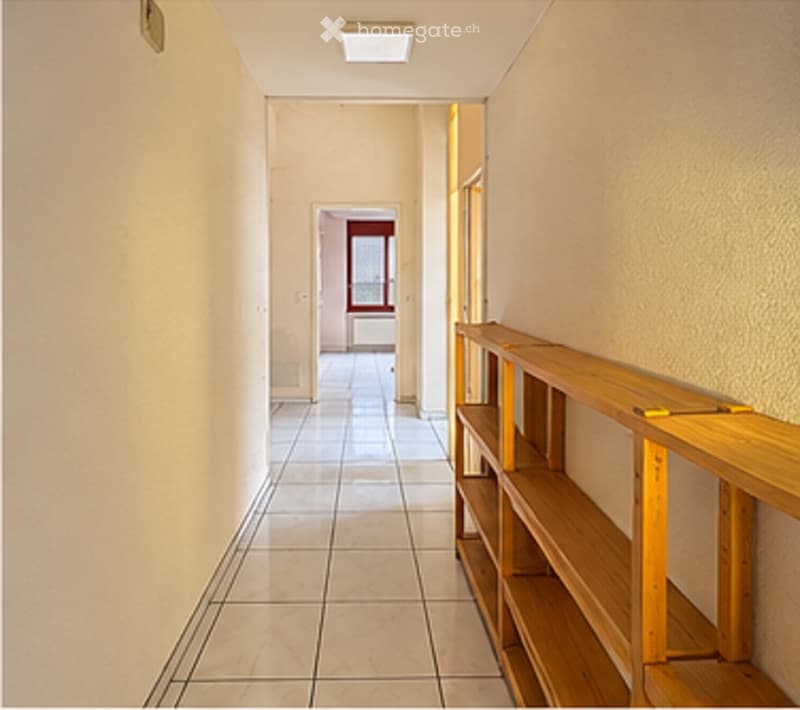 Empty hallway with tiled floor, wooden shelf on the right side, and a door at the end leading to another room.