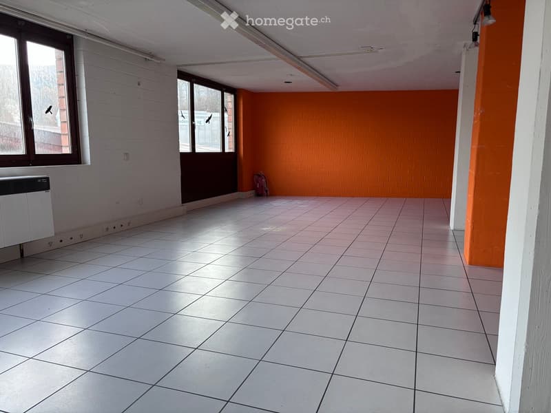 Empty room with white tiled floor, white walls, and large windows. Features include a radiator, a door, and a bright orange accent wall.