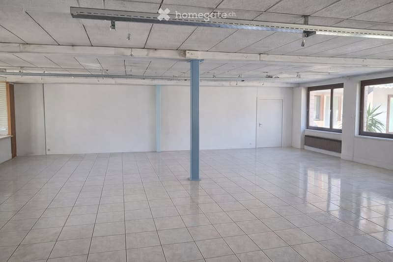 Empty room with tiled floor, white walls, and large windows. Features a central support column and exposed ceiling lighting fixtures.