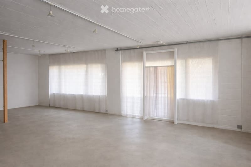 Empty room with white walls, exposed wooden beam, concrete floor, and large windows with sheer curtains.