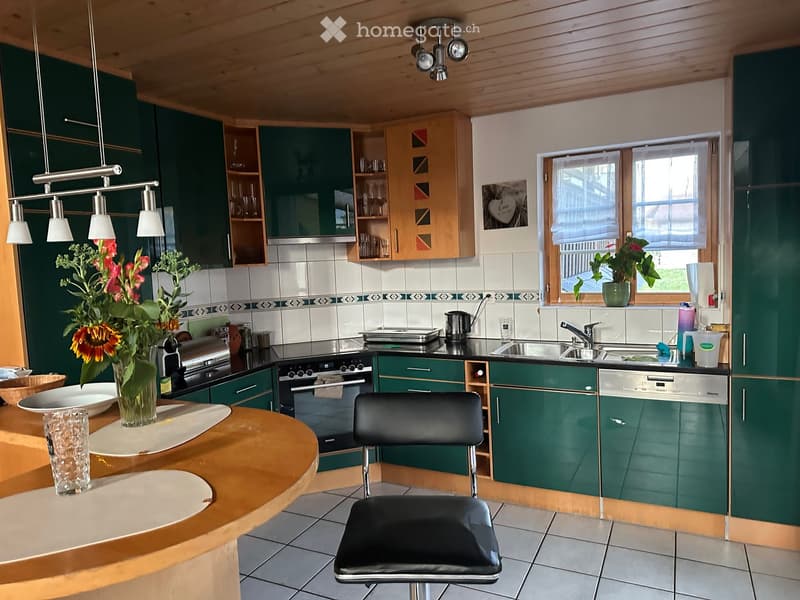 Modern kitchen with green cabinets, stainless steel appliances, a round wooden island, and a black stool. Features include a sink, oven, dishwasher, and decorative vases with flowers.
