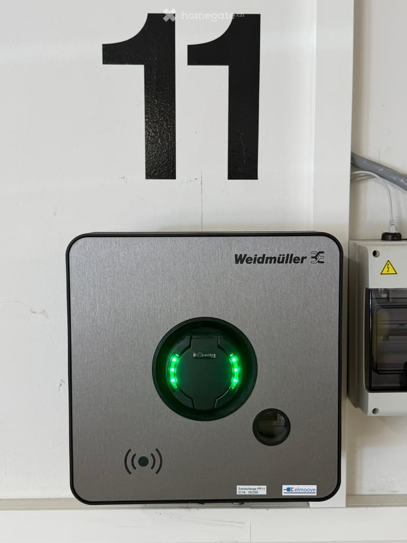 A modern electric vehicle charging station with a sleek metallic finish, featuring a circular charging port with green indicator lights and various certification logos.