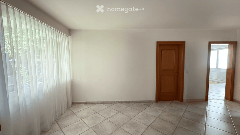 Empty room with tiled floor, white walls, and a wooden door. Natural light filters through sheer curtains on the window.