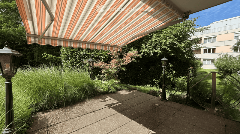 A well-maintained garden area with a striped awning, lush greenery, decorative lampposts, and a paved walkway.