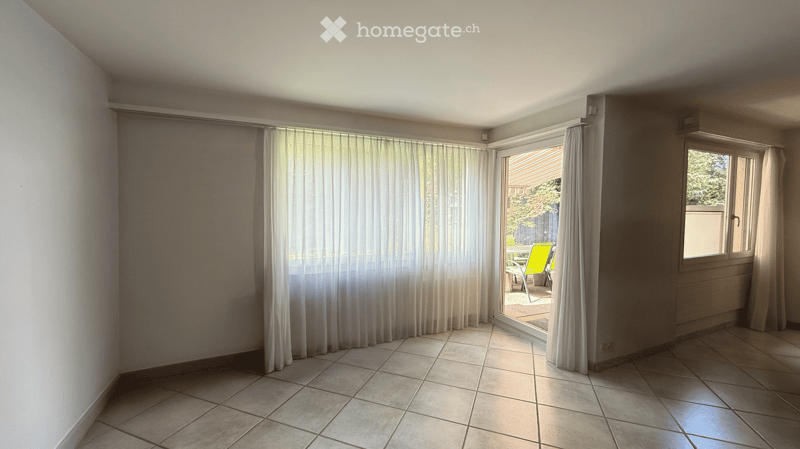 Empty room with tiled floor, large window with white curtains, sliding glass door leading to a patio area with outdoor furniture.