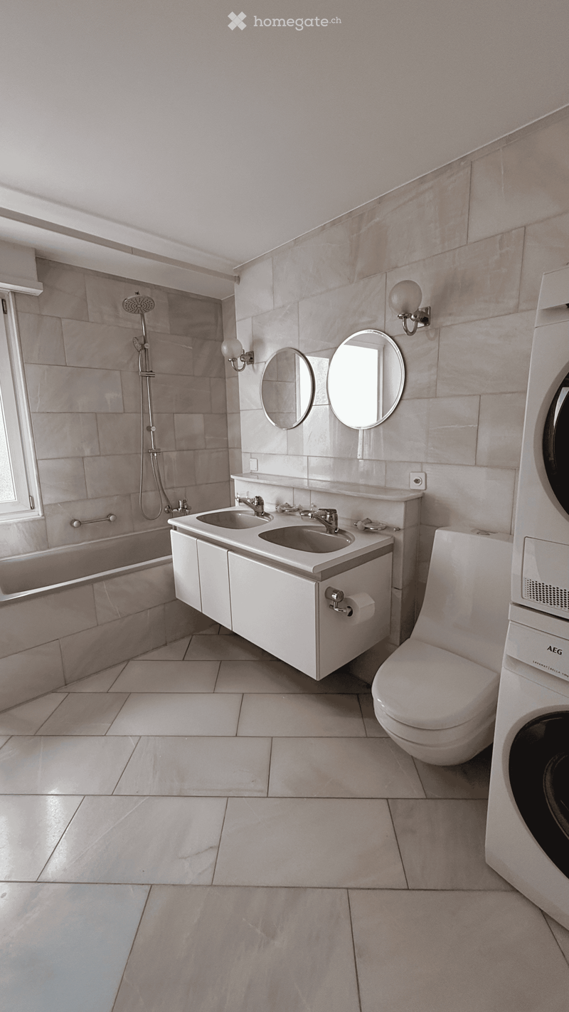 Modern bathroom with beige tiles, double sink vanity, bathtub, toilet, and built-in washing machine. Features two round mirrors, wall-mounted light fixtures, and a shower area with a glass enclosure.